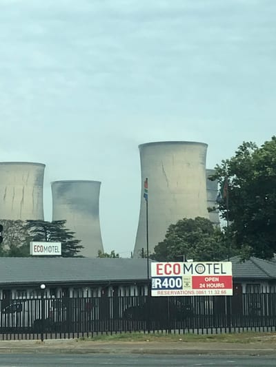 The image depicts an exterior view of the Eco Motel, located at 75 Isando Road, Isando, Kempton Park, South Africa, with large industrial cooling towers visible in the background under an overcast sky.
In the immediate foreground, a dark gray paved road surface is visible, followed by a black metal palisade fence stretching horizontally across the frame. Behind the fence, a single-story motel building, the "ECO MOTEL," occupies the mid-ground. The building features a dark gray tiled roof and light-colored facades with multiple uniform windows and doors. On the roof, to the left, a small white rectangular sign with "ECOMOTEL" in black text is present.
A prominent, large rectangular sign stands directly in front of the motel building. It displays "ECO MOTEL" with a green/black circular logo, followed by "FROM R400" (daily rate), "DAILY" and "HOURLY" options, "OPEN 24 HOURS," and "RESERVATIONS 0861 11 32 66."
To the right of this large sign, a tall black flagpole stands, flying a South African national flag characterized by its black, yellow, green, white, red, and blue colors. Another flagpole is partially visible further back and to the right. Various green trees and shrubbery are scattered around the motel property and behind it.
Dominating the background are three large, light-colored hyperboloid cooling towers, characteristic of a power generation facility. They exhibit vertical streaking and appear weathered. A faint plume of light gray vapor or smoke emanates from the top of the central cooling tower. The sky above is uniformly overcast with light gray cloud cover. No individuals or vehicles are explicitly visible in the scene.