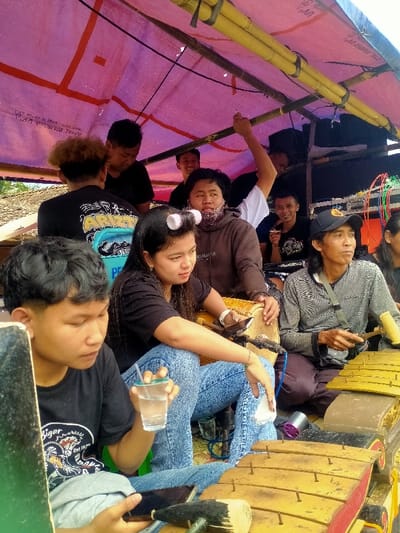 This image depicts a group of approximately nine individuals gathered under a temporary pink tarpaulin canopy, supported by bamboo poles, in Kedawung, Indonesia.
In the foreground, a young male with short dark hair wears a black t-shirt with a white graphic and blue jeans. He holds a clear plastic cup with a straw in his right hand and a black smartphone in his left, looking down.
To his right, a young female with long dark hair, styled with two white hair rollers, wears a black t-shirt and blue jeans. She is seated, with her right hand resting on a light brown kendang drum positioned on her lap. A black smartphone is visible near her left hand. She looks towards the right.
Behind the female, a person in a brown hooded sweatshirt, wearing a patterned face covering, is seated and looks towards the left.
To the right of the masked individual, an older male with dark hair, wearing a black baseball cap and a striped long-sleeve shirt, is seated. He holds two mallets over a gamelan instrument with yellow-brown keys on a red-brown resonator box, and looks towards the right. Another gamelan instrument is partially visible in the bottom right corner. A black microphone is positioned near the kendang drum.
In the background, several other individuals are visible: a male with reddish-brown curly hair in a black t-shirt, another male in a white t-shirt with his right arm raised holding a bamboo pole, and a male in a black t-shirt who appears to be holding a small object to his mouth. The ground beneath the canopy is not clearly visible but suggests a simple, temporary setup.