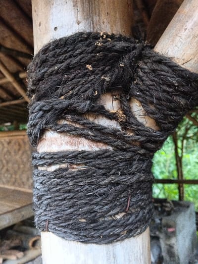 In Kyauk Hsoke, Myanmar, a vertical, light-colored wooden or bamboo post is centrally depicted, tightly bound by multiple wraps of thick, dark, coarse natural fiber rope. The rope exhibits a fibrous, worn texture with scattered lighter, possibly organic, particulate matter. The binding secures the main vertical post to at least one other diagonal wooden or bamboo support, visible entering the frame from the upper right. To the left of the main post, a rustic structure with a woven texture and a thatched roof element is partially visible, alongside a flat wooden surface. To the right, green foliage and additional timber or bamboo structural elements are present against a bright background, with a concrete or stone block partially visible at the lower right. The scene is illuminated by diffused natural light.