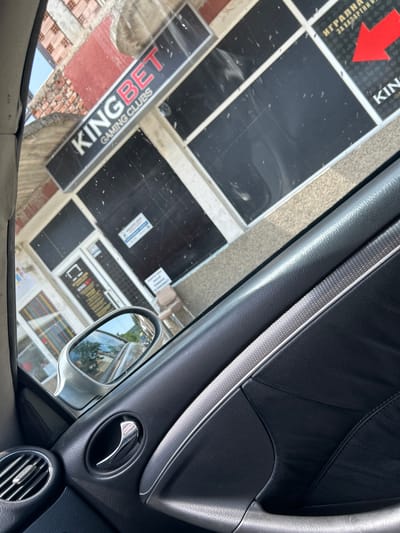 The image, taken from inside a vehicle in Krivodol, Bulgaria, shows a view through the car window of a building with signs for "KINGBET GAMING CLUBS". The building's facade features large black windows and red brickwork. A red arrow points to the right on one of the signs. Inside the car, the focus is on the interior door panel, including a silver door handle, a vent, and textured trim. The side mirror of the car is visible, reflecting trees and the sky. The overall scene captures a street view from a car, focusing on the exterior of a gaming club and the interior of the vehicle.