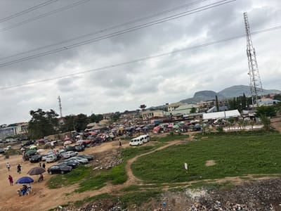 This is a wide-angle, eye-level photograph taken in Angwan Sayawa, Nigeria, depicting a bustling outdoor market scene under a cloudy sky. Power lines stretch across the upper portion of the frame. Dominating the right side of the image is a tall, lattice-style telecommunications tower.

The market itself occupies a large area of dirt and sparse grass. Numerous stalls are set up, many covered by colorful umbrellas and canopies, offering a variety of goods. A significant number of cars are parked throughout the market area, including sedans, SUVs, and several white vans. People are visible walking among the stalls, browsing, and interacting. Some individuals are seated under umbrellas, possibly selling or resting.

In the background, buildings of various sizes and styles are present, suggesting a mixed urban and semi-urban environment. Hills and mountains can be seen in the distance. The foreground of the image shows areas of disturbed earth and piles of refuse, indicating a less developed or maintained section of the area. The overall impression is one of a vibrant, active marketplace in a developing urban landscape.