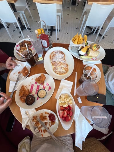A round wooden table sits in a cafe in Miami Beach, Florida, at the coordinates 25.771, -80.134. Three people are seated around the table, and there are several plates of food, glasses, and utensils on the table. One person is holding a fork and knife. Two plates of food are visible in the foreground, containing fried chicken and french toast. A third plate, partially visible, contains a variety of toppings, including syrup, whipped cream, and powdered sugar. Another plate, partially visible, contains a large burrito with a side of chips and salsa. Three glasses of water and a glass of iced coffee are also visible on the table. There are also several napkins, condiments, and paper place mats on the table. The chairs are white and the floor is tiled. The cafe is brightly lit and the atmosphere is casual.