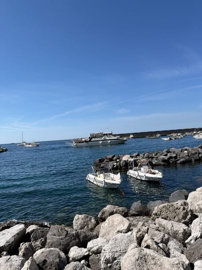 Captured in Pogerola, Italy, the scene features a clear, blue sky streaked with faint airplane contrails above a body of deep blue water. Along the waterfront, a variety of vessels ranging from small boats to larger cruise ships are docked. The foreground consists of a protective barrier of jagged, gray rocks lining the water's edge, with a pair of small white boats nestled amongst them. The scene is bathed in daylight, highlighting the textural contrast between the rough rocks and the smooth water.