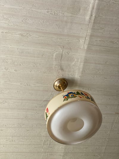 The image shows a close-up view of a ceiling light fixture with a decorative lampshade, mounted on a textured ceiling. The ceiling appears to be paneled with a wood-grain pattern, rendered in a light, neutral color. The light fixture itself has a brass-colored mounting and a cream-colored lampshade adorned with a colorful, whimsical pattern that includes small houses, trees, and figures. The light emanating from the fixture casts shadows and highlights on the ceiling. The image does not provide any information to place it within the city of Чэрвень, Belarus.