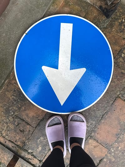 An overhead view captures a detached blue circular traffic sign with a prominent white downward-pointing arrow, resting on an uneven, weathered reddish-brown brick or cobblestone pavement. The sign's surface displays numerous small water droplets, indicating moisture. Positioned directly below the sign are two human feet, clad in light purple slide sandals worn over dark grey socks, with black leggings visible. A grey concrete curb is present along the upper-left edge of the frame. This scene is observed in Cortona, Italy.