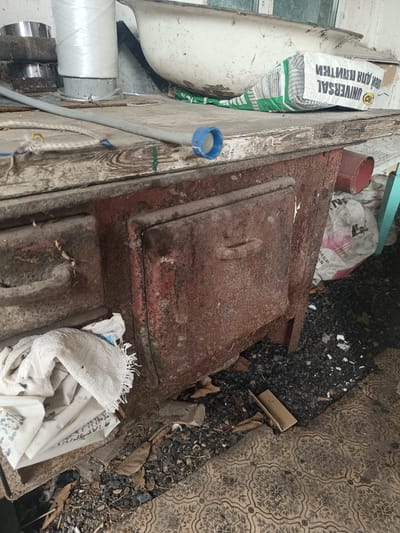 The image depicts an outdoor scene in Ostrogorka, Russia, focusing on a dilapidated, old, rusty cast-iron stove. The stove is the central subject, showing signs of significant wear and tear, with peeling red paint and visible rust. It appears to be a cooking stove, possibly used for heating and preparing food.
To the left of the stove, there are several white woven bags, one of which is partially open and reveals what appear to be more bags or folded material inside. A grey pipe runs horizontally across the top of the stove and a blue cap is visible on its end.
Behind the stove, on a wooden surface, there is a white bathtub. A bag of building material, labeled "UNIVERSAL", is resting on this surface near the bathtub. In the background, more bags and debris are scattered around. The ground beneath the stove and extending to the foreground is covered with small dark stones and some fallen leaves, suggesting an outdoor or semi-outdoor environment. A patterned tiled floor is visible in the lower right corner of the image. The overall impression is one of neglect and disuse. There are no people or animals visible in the image.