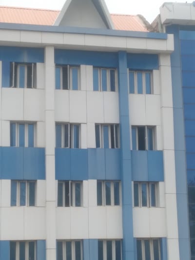 The image shows a section of a modern building in Osu, Nigeria. The building has a white and blue color scheme and a grid-like design, with windows in a regular pattern. The building appears to have multiple floors and is constructed with panels or cladding. Some windows are slightly ajar. The image is taken from a low angle, showing the side of the building. The sky is overcast or bright, and the overall lighting is soft and diffused.