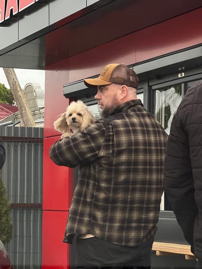 A man wearing a brown and khaki baseball cap and a plaid shirt is holding a small, light-colored dog. The man has a beard and appears to be looking down at the dog. They are positioned in front of a building with red paneling and black trim. The building is located in Velyki Hai, Ukraine.