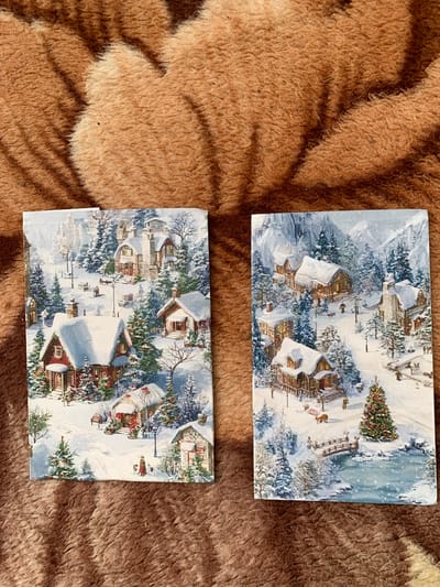 The image shows two rectangular cards placed on a textured brown surface. The cards depict snowy village scenes.
The top card includes images of snow-covered houses and pine trees. A decorated Christmas tree is visible near a partially snow-covered bridge over a stream. There is a suggestion of mountains in the background.
The bottom card also depicts a snowy village scene with buildings and trees covered in snow. There are figures of people and animals in the card.