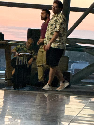 The image shows a group of people in what appears to be a modern airport terminal at sunset. The sky outside the large windows displays a gradient of pink and orange hues. In the foreground, a man wearing a loud, patterned short-sleeved shirt and dark shorts is walking. He has a watch on his left wrist and is wearing white sneakers. To his left, a man with a beard and dark hair wears a dark red t-shirt. Further to the left, a person is seated, wearing a green floral patterned shirt and dark pants. They are holding a dark bag with various items, possibly toiletries or a medical kit, lined up on top. The flooring is reflective, showing the overhead structure and lighting. The overall atmosphere suggests a travel hub.