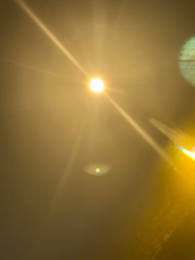 The image shows an intense light source, potentially the sun, obscured by a haze or fog. Rays of light emanate from the central bright spot, creating a starburst effect. A fainter, secondary light spot is visible to the right of the main light source. The overall color palette is dominated by yellow and brown tones. There is insufficient information to establish the location as Unknown, France. The scene appears to be a general environmental condition rather than a specific event involving people or objects.