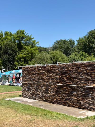 The image depicts an outdoor scene on a sunny day, likely at a recreational area in Simitli, Bulgaria, given the context of the provided crops. In the foreground, a textured stone wall, constructed from horizontally stacked rectangular stones in varying shades of brown and orange, dominates the right side of the frame. Water pipes with visible faucets run along the top of this wall, suggesting it may be part of an outdoor shower or washing facility.
To the left of the stone wall, a patch of concrete is visible, wet in places, indicating recent water usage. Beyond the concrete, green grass covers the ground, transitioning to drier, yellowed grass further into the foreground. A red brick path is partially visible in the middle ground, leading towards a shaded area.
Underneath several blue umbrellas with "Nestle" branding, lounge chairs are set up on the grass. Several people are present in this area. One person, wearing a pink top and yellow shorts, stands near the chairs. Another person in a black swimsuit is also visible. Other individuals are present but less clearly defined. In the background, a dense line of lush green trees forms a natural backdrop, with a hazy outline of hills or mountains visible in the far distance under a clear blue sky. The overall impression is of a summer day at an outdoor facility, possibly a park or a resort, with amenities for cooling off.