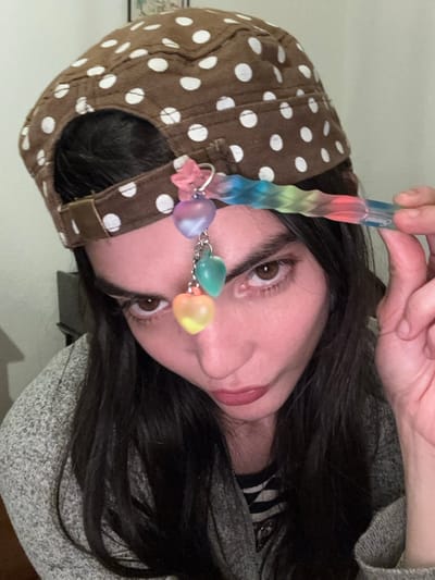 An individual, appearing female, is depicted in a close-up, head-and-shoulders indoor shot. The subject wears a brown military-style cap patterned with white polka dots. Attached to the cap's front-right brim is a decorative charm featuring a pink butterfly figure connected to a metallic ring, from which a chain suspends three translucent heart-shaped beads: the uppermost is purple/pink, the middle is green/teal, and the lowest is yellow/orange. The subject's right hand holds a multi-colored, translucent, twisted object, exhibiting a gradient of blue, green, yellow, orange, and pink, positioned near the right temple. The subject has dark hair, prominent eyebrows, and brown eyes with visible eyeliner, gazing directly forward with pursed lips. A light grey, textured cardigan covers a striped garment. The background is an indistinct, monochromatic indoor environment, offering no discernible features to specify the exact location within Roanne, France.