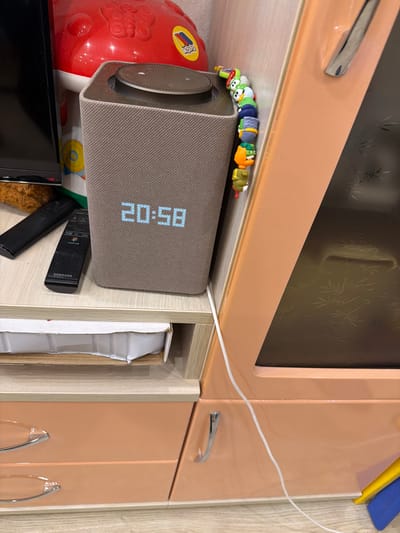 The image shows a close-up view of a modern speaker with a digital display showing the time "20:58". The speaker is on a wooden shelf, to the left of a large, orange piece of furniture with a reflective surface. A string of small, colorful cartoon figures is draped over the speaker. To the left of the speaker, there are two black remote controls. Behind the speaker, a red plastic toy with a butterfly design is visible. The background includes a wooden floor and the side of a cabinet. The scene appears to be indoors, in a domestic setting, possibly a living room or bedroom. The image does not provide specific geographical information for Александровка, Russia.