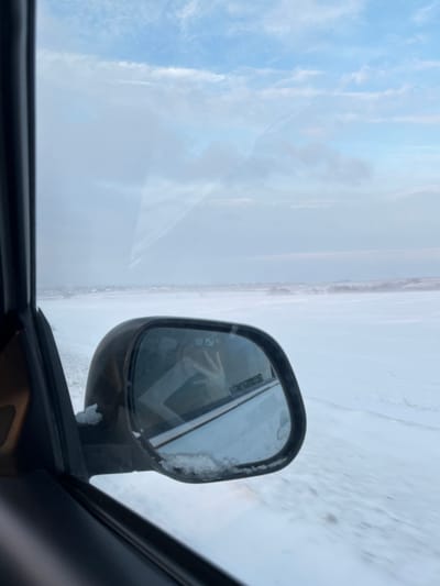The image depicts a winter scene captured from the interior of a vehicle in Gostagaevskaya, Russia. The view primarily features a snow-covered landscape under a partly cloudy sky with light blue and grey tones. On the left, a dark-colored vehicle door frame and window pillar are visible. A black-framed side-view mirror, partially encrusted with snow and ice along its lower edge, is prominent in the mid-left. Reflected within this mirror is a person's hand, holding a white or light-colored smartphone, indicative of the photographer. The reflection also shows a portion of the vehicle's white exterior. Beyond the vehicle, a vast expanse of snow covers the ground, with faint tire tracks or a road visible to the immediate left. In the distance, a cluster of structures, likely buildings, marks the horizon, indicating the edge of the settlement. Further right, a line of snow-dusted vegetation or trees can be observed. The overall environment is characterized by cold, snowy conditions.