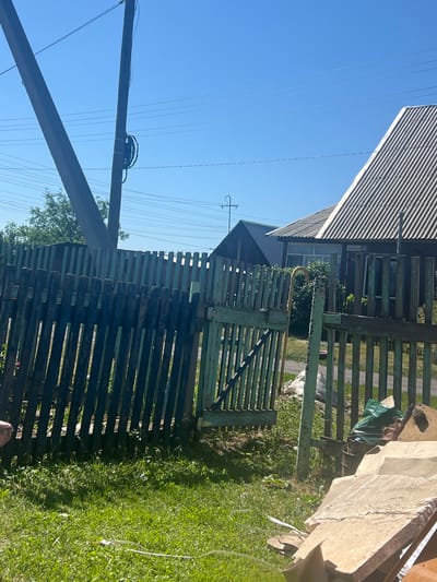 The image depicts a sunny outdoor scene in Zudilovo, Russia. In the foreground, a weathered wooden fence, painted a faded teal, runs diagonally across the frame. A gate within the fence is ajar, revealing a glimpse of a grassy area beyond. To the right of the fence, a pile of construction debris, including large flat pieces of what appears to be concrete or stone, is visible. A green tarp and a dark barrel are also present amidst the debris.
In the midground, the fence continues, bordering a property with a dark wooden house and a house with a corrugated metal roof. A street sign with Cyrillic lettering and the number "45" is affixed to the side of the house with the metal roof. A tall wooden utility pole with electrical wires stretching across the sky stands prominently on the left side of the image. Trees and foliage are visible behind the fence, adding to the rural or suburban character of the location. The sky is a clear, bright blue. No people are visible in the frame.