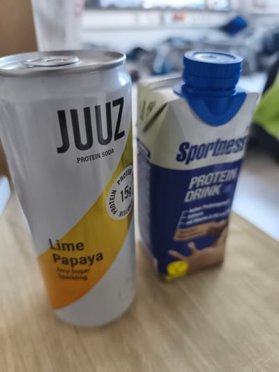 Two beverage containers are on a wooden surface. To the left is a white aluminum can with the brand name "JUUZ" in black lettering. Below "JUUZ" it says "PROTEIN SODA". A yellow and orange gradient is visible on the lower part of the can, with the text "Lime Papaya Zero Sugar Sparkling" printed on it. A circular label on the can indicates "15g PROTEIN". To the right is a white cardboard carton with a blue cap. The carton displays the brand name "Sportness" in blue lettering and "PROTEIN DRINK" below it. A dark brown liquid with milk splashing out of it is depicted on the front of the carton.

The location in Troisdorf, Germany is not identifiable from the provided image.
