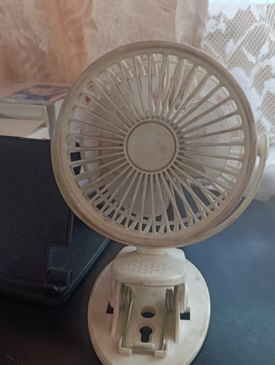 A close-up shot focuses on a small, white, clip-on fan. The fan is positioned on a dark surface, likely a desk or table, with a black object, possibly a speaker, to its left. In the background, sheer curtains, one with a lace pattern, are visible. The fan itself appears to be dusty, with a weathered appearance. The clip mechanism at the base of the fan is prominent, featuring silver-colored springs and a hole for attachment. The overall scene is indoors, with soft lighting that creates subtle shadows. There is no apparent activity or event taking place; the image seems to be a still life showcasing the fan. The location cues are limited to the curtains and the general indoor setting. No visible text is present in the image. The time of day and weather are not discernible.
