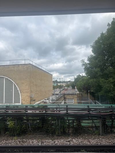 The image depicts an urban landscape on a cloudy day. In the foreground, a railway track is visible, characterized by ballast stones and a black metal rail. Above the tracks, a complex network of dark pipes and cables is supported by metal structures, with some sections covered in green ivy.
Further back, a light-yellow brick building with a distinctive arched window featuring a grid pattern occupies the left side of the frame. A satellite dish is mounted on the side of this building. Beyond the building, a road stretches into the distance, lined with streetlights and trees. A red bus is seen traveling on the road. The background features more buildings and lush green trees, suggesting a mixed urban and suburban environment. The sky is overcast with heavy grey clouds. The location is identified as Epping Forest, United Kingdom.