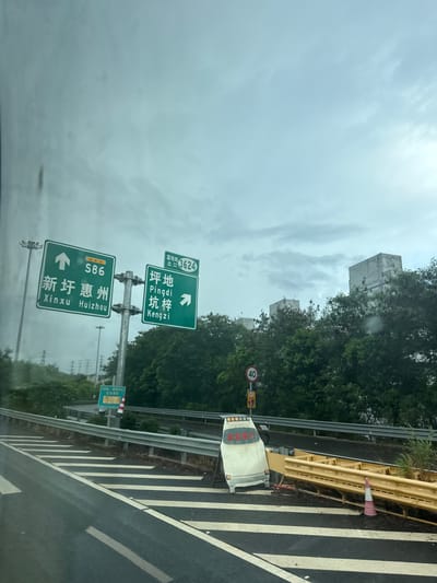 The image shows a highway interchange in Longgang District, China, under a cloudy sky. Green directional signs indicate that S86 leads to Xinxu Huizhou and that Pingdi Kengzi is straight ahead, with an exit number 3624. A damaged white vehicle, possibly a utility cart, is partially visible near a bent yellow guardrail. A speed limit sign indicates 40 km/h. In the background, trees and modern buildings are visible. The road surface shows white lane markings and some debris.