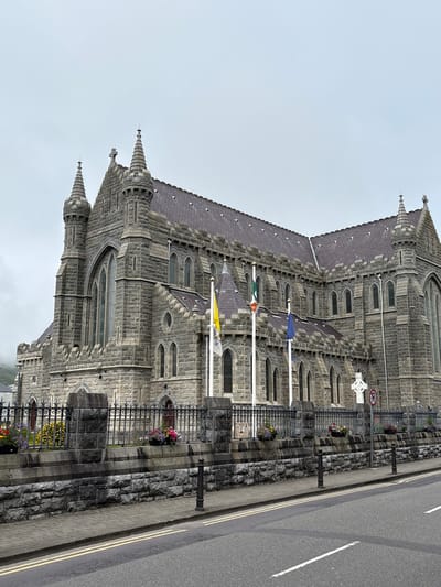 The image depicts the exterior of a large, gothic-style stone cathedral located in Cahersiveen, Ireland. The cathedral features prominent towers with conical spires and numerous arched windows. A wrought-iron fence encloses a garden area in front of the cathedral, where colorful flowers are planted in decorative baskets. Several flags, including the Irish tricolor, are visible on flagpoles in front of the building. A white Celtic cross stands to the right of the flags, along with a red and white circular sign indicating "no parking." The cathedral is situated adjacent to a paved road with double yellow lines. The sky is overcast and the overall atmosphere is somber.