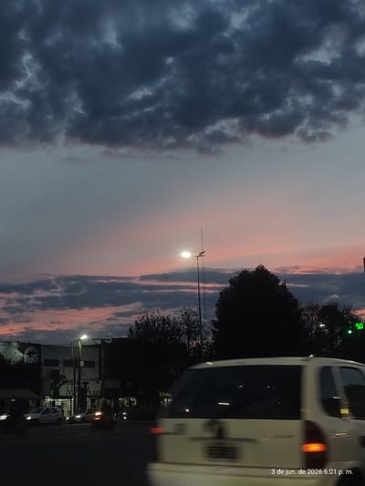 The image depicts an urban street scene at twilight, captured at 6:01 p.m. on June 3, 2026, in Sarandí, Argentina. The sky dominates the upper portion of the frame, showcasing a striking contrast of dark, heavy clouds on the left, which gradually transition into softer pink and orange tones of a sunset towards the right.

In the middle ground, a bright streetlamp illuminates the scene, its glow prominent against the fading daylight. Silhouettes of trees, likely on the roadside, stand out against the horizon. On the right side of the image, a white vehicle, possibly a van or bus, is partially visible and appears to be in motion or parked on the street, with its red taillight glowing.

Further down the street, buildings with illuminated windows suggest commercial activity. Visible text on these buildings includes "Expre" and "SHOP," indicating businesses, along with fragments like "Ricat" and "M." A green illuminated "50" is also discernable in the upper right, which could be part of a traffic sign or a digital display. The overall impression is an atmospheric evening in a city, where artificial lights begin to complement the last vestiges of daylight.