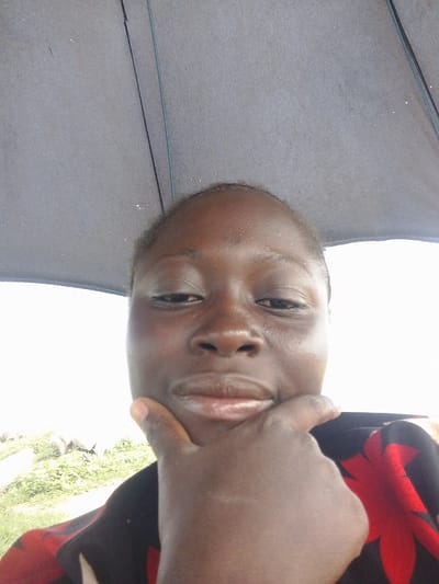 A young African person is taking a selfie from under a dark umbrella. The person's chin is resting on their hand, and they are wearing a black and red patterned shirt. The background is blurred and appears to be an outdoor scene with greenery and possibly some rocks. The image is taken at a close-up perspective, focusing on the person's face. The location is identified as Gosa, Nigeria.