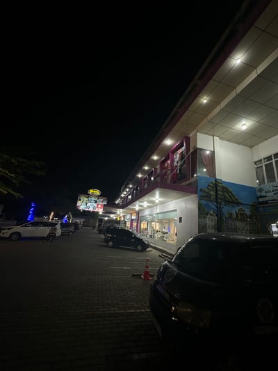 The image depicts a nighttime scene in front of a commercial building complex in Keudeteupinpunti, Indonesia. Several cars are parked in a paved lot. The building has multiple levels with illuminated windows and signage. A large, brightly lit sign for "SUMBER" is visible, along with other advertisements and murals on the building's facade. The environment is dark, with artificial lighting from the building and streetlights illuminating the scene. There is a traffic cone near the foreground.
