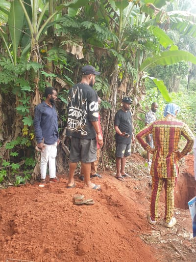 Several people stand near a mound of red earth in what appears to be a forested area. The location is Nkwerre, Nigeria. The individuals are dressed in casual clothing. One individual wears a colorful patterned suit. The ground is covered in red soil and vegetation. A clump of banana trees are growing behind the people.