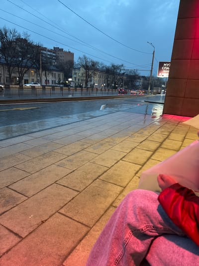 The image depicts a street scene in Ol'ginskaya, Russia, on an overcast day. In the foreground, a person wearing jeans and a red jacket is holding a bouquet. The ground is paved with rectangular stones. Part of a building with a dark-colored facade is visible on the left, with a street running alongside it. A sign reading "МУ) СПА. (БАНІ" is also visible. In the background, there are apartment buildings, bare trees, and a row of cars on the street. The street surface appears wet, possibly from rain.
