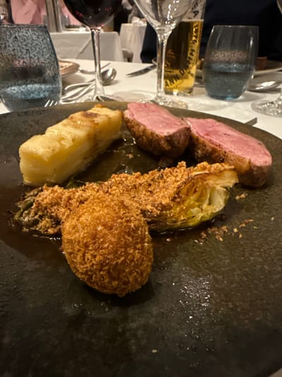 The image captures a plate of food, seemingly taken at a restaurant. The primary elements are slices of what appears to be rare duck or lamb, accompanied by fried, breaded Brussels sprouts, and a soft white vegetable. The plating is rustic, on a dark grey or black textured plate. In the background, the dining table setting is visible, including wine glasses, water glasses, cutlery, and a bottle of alcohol, likely wine or whisky. Due to the presence of a bottle labeled "1876" it might be related to a special occasion. The scene is indoors, likely a restaurant. Without further context, the location is unknown, and I can not relate to Glenridding, United Kingdom.