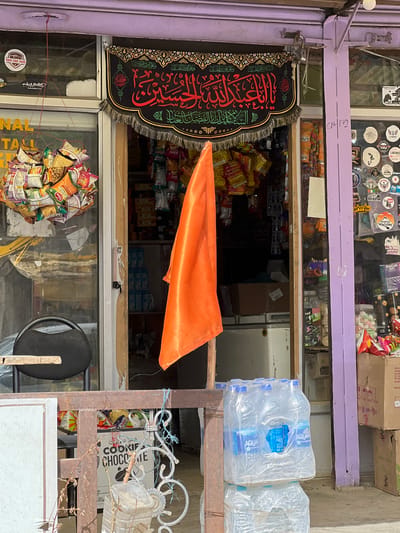 The image depicts the exterior of a small shop in Yogma Mangmore, India. The shop entrance is adorned with a black banner featuring Arabic calligraphy in red and green, with a fringed bottom. An orange flag is prominently displayed hanging from an orange pole, partially obscuring the shop's interior. The shop appears to sell snacks and drinks, with various packets of snacks visible hanging from the ceiling and shelves inside. To the left of the entrance, a large bunch of snack packets is suspended by a string. A black chair is visible through the glass door. The shop front is decorated with numerous stickers on its purple-painted pillar. In the foreground, a stack of bottled water is placed outside the entrance, and a metal railing separates the entrance from the viewer. The ground is paved with concrete.
