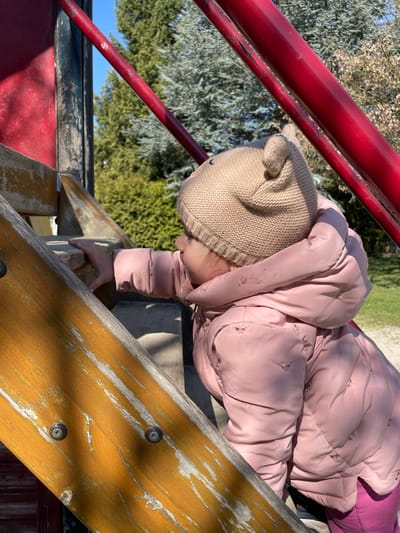A young child is depicted outdoors on a sunny day, actively climbing a playground structure. The child, viewed in profile from the mid-torso up, wears a light brown knitted beanie with two small ear-like protrusions and a light pink puffer jacket featuring subtle embroidered patterns. A reddish stain is present on the upper left shoulder area of the jacket. The child's left hand grips a worn, light brown wooden step, which exhibits peeling paint or varnish and exposed metal screws. Reddish-orange painted metal bars, showing minor abrasions, are visible above and around the child, forming part of the playground equipment. The background consists of various green trees, including coniferous types, under a clear blue sky. A light-colored ground surface, consistent with gravel or dirt, is visible behind the child. While the setting is within Senden, Germany, the specific location or address within the city cannot be determined from the visual information provided.