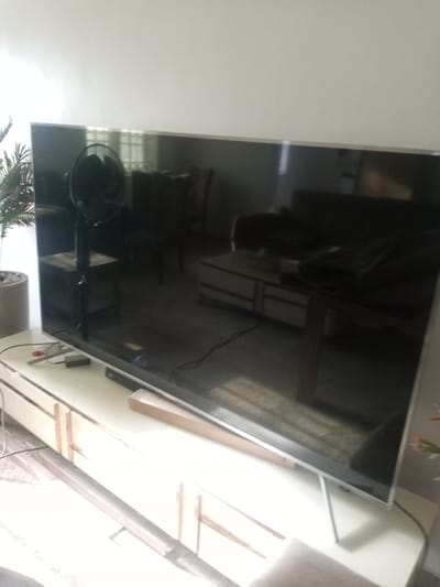 The image shows a large, modern television screen dominating the center, reflecting the room. To the left, a tall, cylindrical planter with a plant is visible, partially obscured by the TV. Beneath the television, a long, white media console with drawers is present, with some cables and electronic devices on its surface. In the reflection on the TV screen, a fan, a coffee table with drawers, and furniture like a sofa are discernible, suggesting a living room environment. The overall scene is a well-lit indoor space, likely a home in Hotoro North, Nigeria.