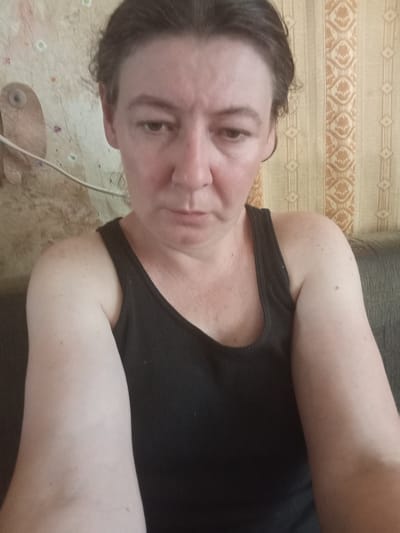 A woman is pictured from the chest up, taking a selfie. She is wearing a black tank top. Her hair is dark brown and pulled back. The background appears to be a room with patterned wallpaper in shades of yellow and beige. To the left of the woman, there is a light-colored object on the wall that resembles a bird or a statue, with a wire running past it. To the right, a dark-colored sofa is visible. The lighting suggests an indoor setting, possibly during the day, with no specific indicators of the city of Berezivka, Ukraine.