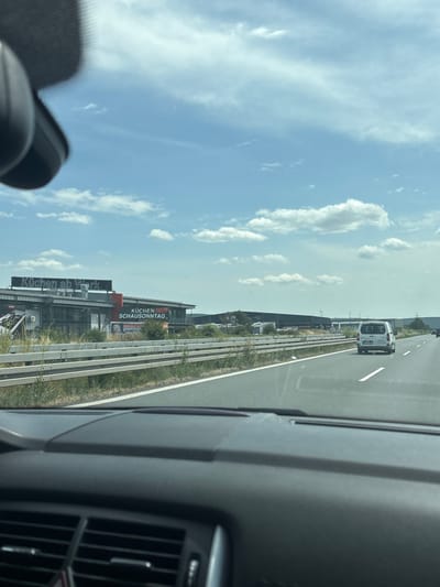 The image captures a view from inside a vehicle traveling on a highway. The foreground shows the dashboard and the lower portion of the windshield. Beyond the highway guardrail, a commercial building is visible with signs reading "Küchen ab Werk" and "KÜCHEN SCHAUSONNTAG". Further in the distance, several other industrial buildings and a white van are seen on the highway. The sky is blue with scattered clouds. This location is the city of Großwallstadt, Germany.