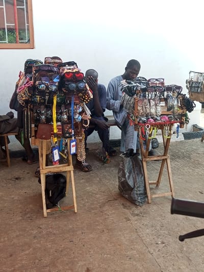Two men sit behind stands displaying sunglasses, wallets, and trinkets. One man wears a light blue long-sleeved shirt and is looking at the sunglasses on his stand. The other man wears a dark blue long-sleeved shirt and has his hand to his face. The stands are simple wooden structures. A black bag rests beneath the stand on the left, and a gray trash bag sits beside the stand on the right. A window with a red frame is visible in the background.