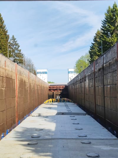 The image shows the interior of a river lock, most likely located in Alekseevskoe, Russia. The lock is constructed of large, rectangular concrete blocks forming the walls. A barge is positioned inside the lock. Several people are visible on the deck of the barge. Overhead, a red bridge spans the lock. The sky is blue with scattered clouds. Tall evergreen trees line the sides of the lock. Streetlights are interspersed among the trees. The overall environment suggests a sunny day at a working waterway infrastructure facility.