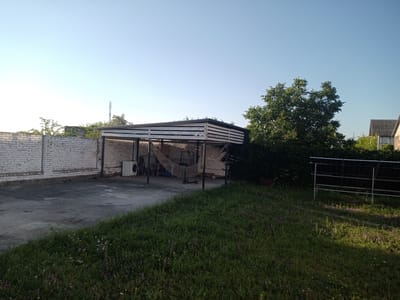 The image depicts an outdoor scene in Blizhnii Khutor, Moldova. A white brick wall runs along the left side of the frame, bordering a concrete area. In the center, a carport with a slatted roof is visible, supported by metal poles. Inside the carport, a white washing machine, a hammock, and a kick scooter are present. A large tree with green foliage stands to the right of the carport, partially obscuring two buildings with pitched roofs in the background. In the foreground, a grassy area with scattered wildflowers and a solar panel array are visible. The sky is clear and blue. There are no people or animals present in the image.