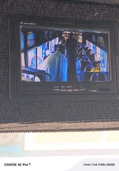 A small black TFT color monitor displaying a scene inside a bus. People are seated and standing within the vehicle, visible through the bus windows and interior. The monitor has buttons labeled "V1/V2," "MENU," "+," "-", and a power symbol along the bottom. The monitor is mounted on a dark gray, textured surface. The bottom of the image reveals a white surface. In the lower section, text states "CAMON 40 Pro" alongside camera settings "71mm f/1.8 1/100s ISO105."