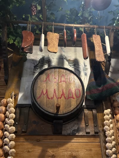 The image depicts a rustic interior scene with a distinct European or old-world aesthetic. The primary focus is a wooden barrel, possibly a wine or liquor barrel, mounted on a wooden wall. Red lettering is painted across the barrel's surface. The wall is adorned with strings of garlic and onions, adding to the old-world atmosphere.
Hanging to the left of the barrel is a display of various meats and sausages, strung up and suspended from a wooden frame. Faux vines and grapes enhance the setting, suggesting a cellar or tavern-like environment. The overall lighting is dim, accentuating the rustic and possibly historical theme.
There are no people visible in the frame, and no specific actions or interactions are depicted. The location is not directly evident from the image, and it is impossible to confirm the city of Des Plaines, United States of America, as the location based solely on this visual information.
