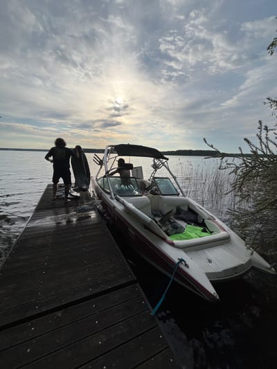 Scene: A partially-overexposed outdoor scene at a lakeside location, possibly near Пятница, Russia, although definitive proof is lacking.
Subjects: Two people are visible on a wooden dock. One individual, seen from the rear, stands towards the edge of the dock wearing a dark t-shirt and shorts. Another person is visible inside a boat tied to the dock.
Objects: A white boat with a red stripe is moored alongside the dock. The boat contains what appears to be a life jacket and other items. The dock itself is constructed of dark-colored wooden planks. To the side of the boat are reeds or tall grass, indicating a natural shoreline.
Actions: One person is standing on the dock. The person in the boat appears to be engaged in some activity inside the boat.
Interactions: It is unclear if the people are interacting directly, but their proximity suggests they are connected.
Environment: The scene is set on a lake with a distant treeline visible on the opposite shore. The sky is bright with scattered clouds and the sun breaking through, causing the overexposure in parts of the image. The water is calm.