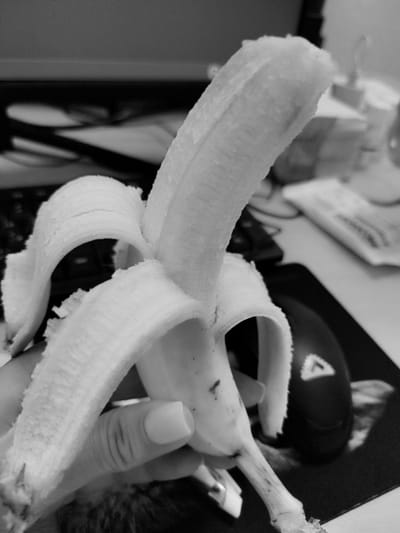 A partially peeled banana is held in a hand in the foreground. The banana is positioned vertically, with its peeled section curving upwards and to the right. The background is blurred, but appears to be a desk with a keyboard, a computer mouse, and possibly some papers. The image is in black and white. There is no indication of the city of Skadovsk, Ukraine, or any specific urban environment.