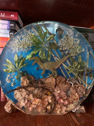 This media file showcases a circular, transparent resin art piece embedded with various natural elements and miniature figures. The scene depicts a prehistoric or fantasy landscape. A green dinosaur figure, possibly a velociraptor, is positioned centrally, as if stalking through lush vegetation. Surrounding it are dried flowers in shades of pink and beige, along with rocks and what appear to be twigs or dried plant stems, all suspended in a vibrant blue, glittery resin. In the upper right portion of the scene, a small turtle figurine is visible amongst the rocks. The overall impression is a miniature diorama encased in resin.
The resin piece rests on a stack of books, with their spines partially visible. The text on the books, some in Cyrillic script, includes titles such as "Ы," "Горам," "етром св," and "Солни." There is also visible text in what appears to be a European language, possibly Slovenian, related to a "laser" device, mentioning "genialen," "lasersko linijo," and "samoniveliren." The background suggests an indoor setting with wooden surfaces, likely a shelf or table. The lighting appears to be artificial, casting some reflections on the resin. There are no people or specific weather or time-of-day cues evident. The Izborsk, Russia location context is not visually discernible from the image itself.