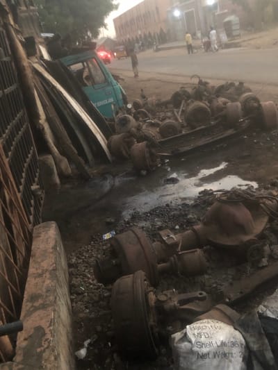 The image depicts an outdoor scene, likely a scrap yard or a large mechanic's workshop, filled with various discarded vehicle parts. The image is oriented vertically (portrait mode), but the scene itself is best interpreted in landscape mode, with the main subject matter spread across the ground.
In the foreground, a large, rusty metal grate or drainage cover is partially visible on the right. To its left, a thick, rectangular concrete block or slab sits on the ground. The ground itself is uneven, composed of dirt, gravel, and several puddles or damp, muddy patches.
The midground is dominated by a dense collection of heavy vehicle components, primarily what appear to be truck axles, differential assemblies, and large brake drums or wheel hubs. These parts are heavily soiled, rusted, and arranged in disarray, suggesting they are salvaged or awaiting disposal/repair. Some appear to be connected by wires or cables. On the far right side of this collection, a crumpled white sack or bag is visible, with some printed text. Part of the text that can be read includes "Batch No.", "MFD", "Shelf Life: 36", and "Net Weight:". A small, colorful wrapper is also on the ground near these parts.
In the background, to the left of the main pile of parts, a light-colored, possibly teal or green, truck is partially visible. Its rear brake lights are illuminated red, suggesting it might be stopped or idling. On the side of the truck, some faded white text is discernible, including "SERVICE JOBS" and a number that looks like "208" or similar. Further into the background, above and behind the truck, there is a building or a sheltered area with several bright artificial lights visible, creating a soft glow in the otherwise dim environment. Several indistinct human figures are also visible in the distance near this structure, one appearing to wear a yellow top.
The overall lighting suggests it is either late evening, dusk, or early morning, with artificial lights playing a significant role in illuminating the scene. There is no specific activity clearly taking place; the scene appears to capture a static view of a storage or work area.