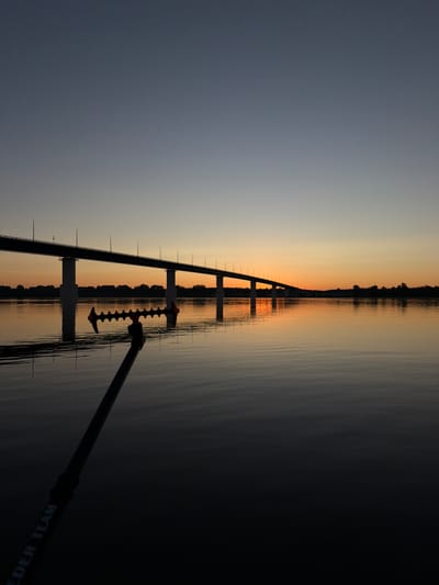 A wide shot captures a tranquil river scene at sunset, featuring a long bridge spanning across the water. The sky displays a gradient of colors, from a deep blue at the top to warm orange and yellow hues near the horizon. The bridge, supported by several concrete pillars, has a railing and a series of lampposts running along its length. The water's surface is calm, reflecting the bridge and the colorful sky, creating a mirror-like effect. In the foreground, a portion of a fishing rod with a specialized rest is visible, angled towards the water. The rest has an orange and black design with multiple indentations. The rod itself is dark and has white lettering that reads "FEIDER TEAM". The background shows a silhouetted treeline on the far bank of the river, indicating land. The overall atmosphere is peaceful and serene, suggesting a quiet moment by the water during twilight.