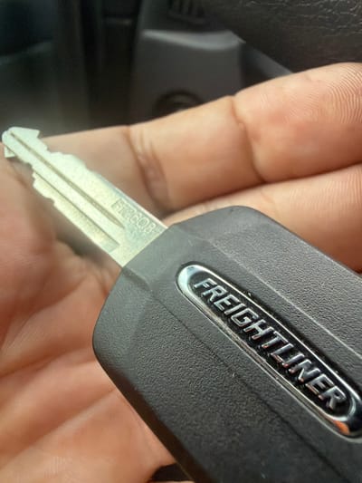 A close-up, eye-level shot shows a person's hand holding a black Freightliner key. The key's blade is silver and has a jagged edge. Embossed on the black plastic part of the key is the word "FREIGHTLINER" in silver lettering. The background is dark and out of focus, with hints of a vehicle interior visible. The image was taken in Murfreesboro, United States of America.