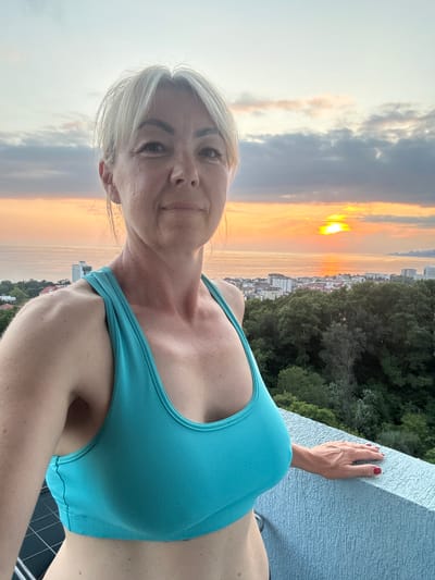 This is a selfie of a woman taken from a high vantage point, possibly a balcony or rooftop, overlooking a city at sunset. The woman has blonde hair and is wearing a turquoise sports bra. Her right hand, with red nail polish, is resting on a light blue textured surface that appears to be a railing or wall.

The background displays a dramatic sunset with clouds reflecting shades of orange, yellow, and gray. Below the sky, a city with buildings and dense green trees is visible, suggesting an urban setting surrounded by nature. The time of day is clearly sunset, indicated by the warm hues in the sky and the position of the sun. The weather appears to be clear enough for a sunset view.

The woman is looking directly at the camera with a slight smile, appearing relaxed and content. The location is identified as Adlerskiy, Russia. There is no visible text in the image.