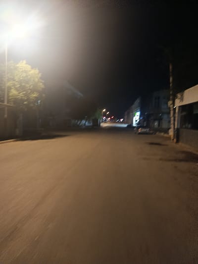 The image depicts a street scene at night in Sumgayit, Azerbaijan. A bright streetlamp illuminates the left side of the frame, casting a strong glow on a tree with green foliage. The street itself is paved and appears empty, with a slight blur suggesting motion, possibly from a moving vehicle. On the right side of the street, several buildings are visible, some with illuminated windows. One building has a sign with green lettering, possibly a shop or business. A car is parked on the right side of the street. The overall atmosphere is dark and somewhat deserted, characteristic of a quiet urban area at night.