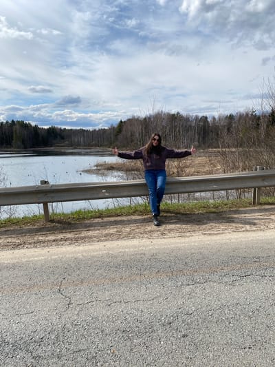 A young woman stands leaning against a guardrail on the side of a road. She has long brown hair and wears sunglasses, a dark jacket, and blue jeans. She has her arms outstretched. To her left, beyond the guardrail, is a body of water. On the opposite side of the water is a forest. The sky is overcast. The road appears to be paved with gray asphalt. The location is potentially Kamennyi Kliuch, Russia, judging by the question prompt.